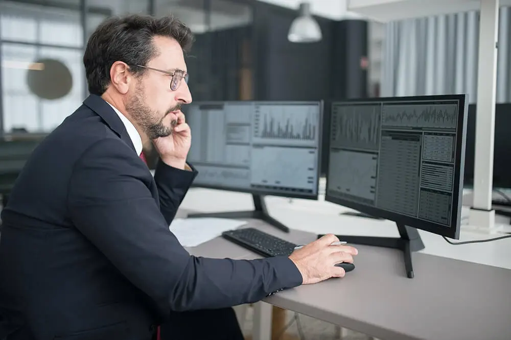 A man wearing a suit looks at graphs on computer monitors.