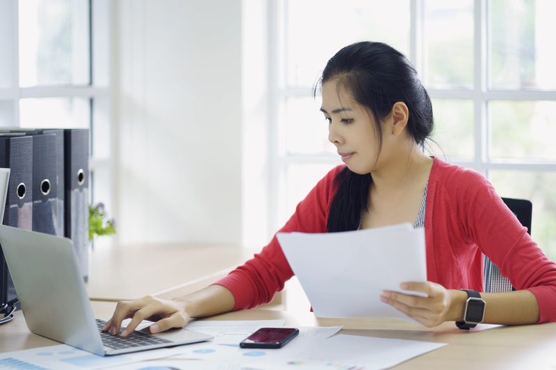 A woman in a red shirt holds papers and looks at a laptop.