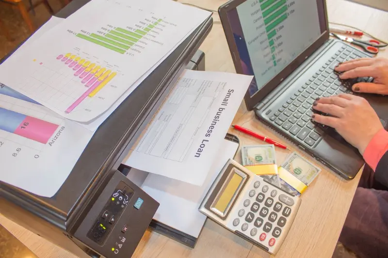 A person works at a desk with a laptop, printer, and multiple pages of charts.